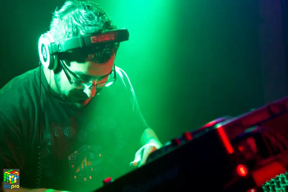 MontsDe in glasses and headphones, lit green, mixing at a DJ event