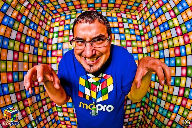 MontsDe in a blue Mo'Pro cube-logo shirt, grinning into a fisheye lens inside a room tiled with colorful squares