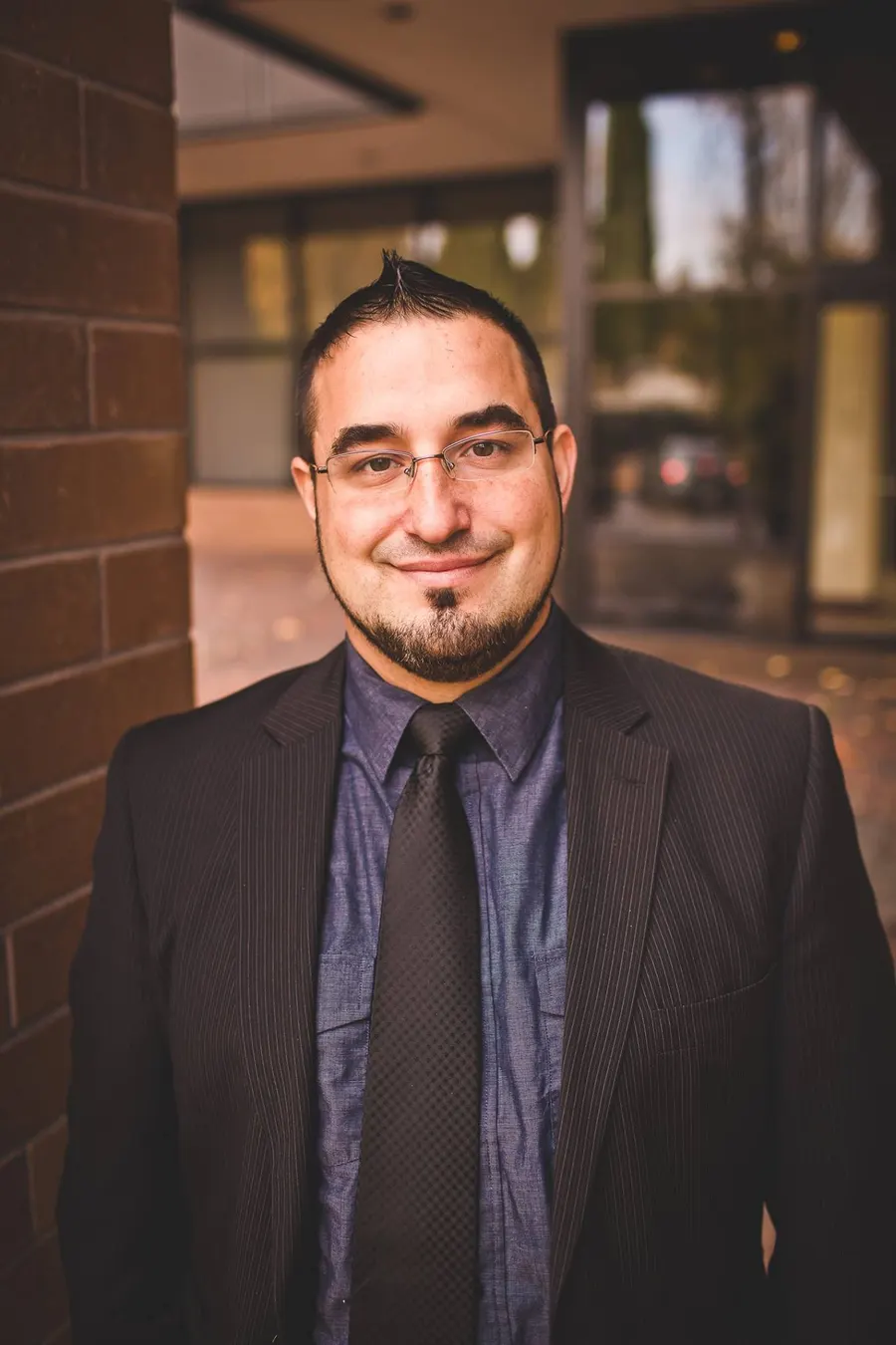 Portrait of MontsDe in a dark suit and tie, smiling, with glasses and a goatee