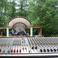 Large analog mixing console in front of the bandshell stage in the trees