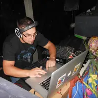 DJ working on a laptop beside mixing gear at night with foliage behind