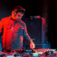 DJ in a red jacket mixing on a laptop and controller under red stage light