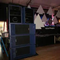 Stacked blue speaker cabinets beside a DJ table in a dim hall with white paper-cut triangles as decor