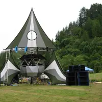 Wide view of a peaked tent stage in a field with speaker stacks and forested hills
