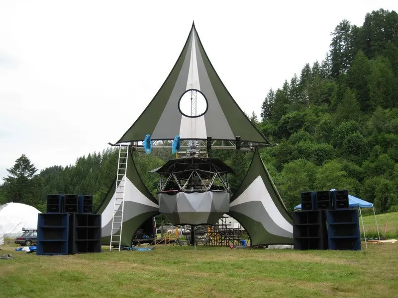 Peaked tent stage in a field with stacks of speaker cabinets beside it and forested hills behind