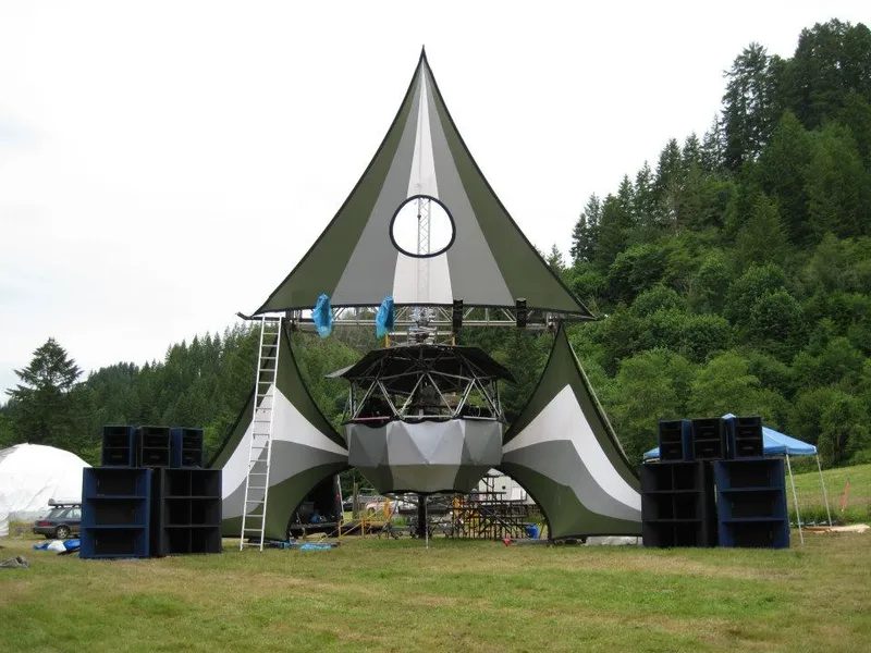 Mo'Pro Crew horn-loaded speaker stacks either side of an outdoor festival stage with a sail-shaped canopy, in front of a forested hillside